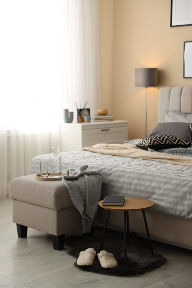 Comfortable bed with soft linens, slippers, pouf and side table in room. Home textile